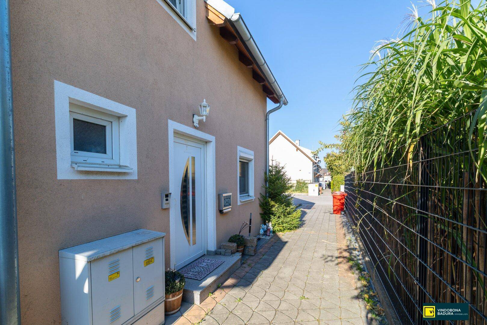 Seitlicher Eingangsbereich des Hauses mit gepflastertem Weg und hohen Pflanzen als Sichtschutz.