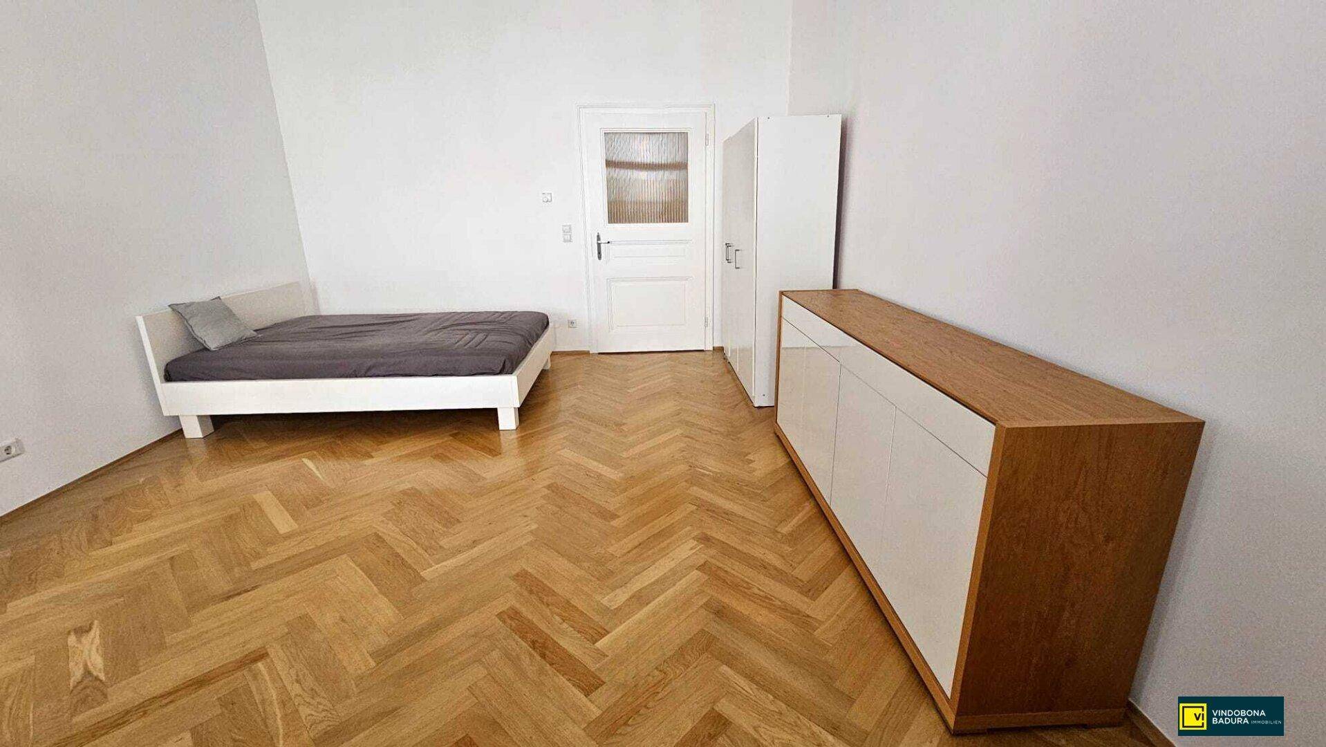 Geräumiges Schlafzimmer mit Fischgrätparkett, einem Bett und einem Sideboard.