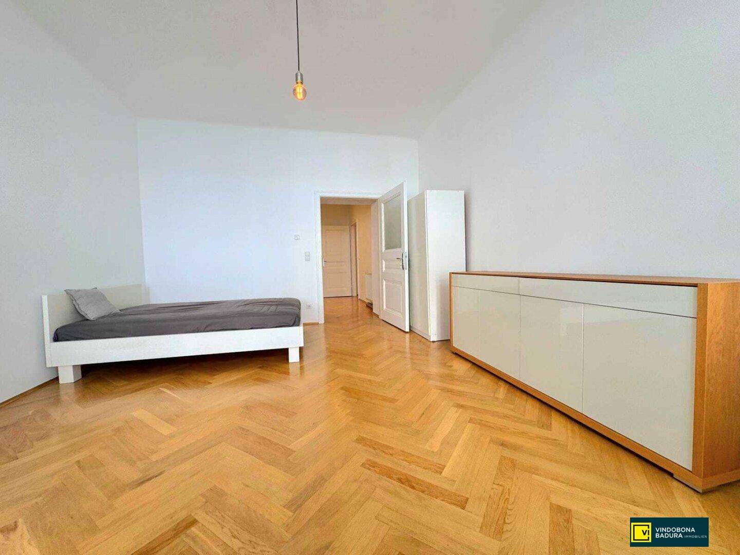 Helles Schlafzimmer mit Fischgrätparkett, einem Bett und einem Sideboard.