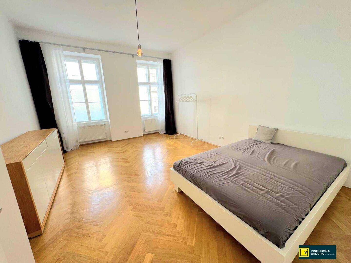 Geräumiges Schlafzimmer mit Fischgrätparkett, großen Fenstern und einem Bett.