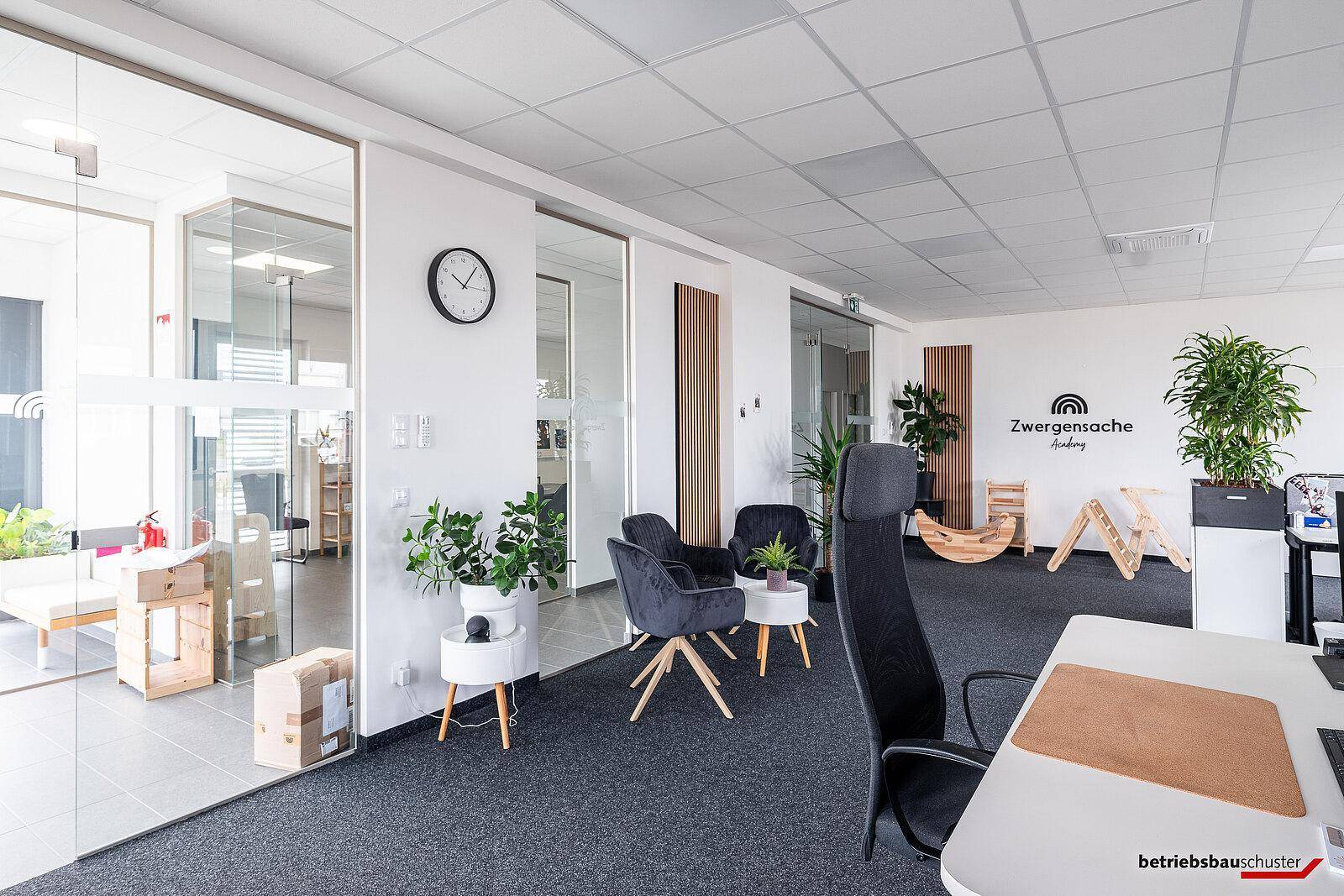 Offener Bürobereich mit Teppichboden, modernen Sitzgelegenheiten und Glaswänden zu angrenzenden Räumen.