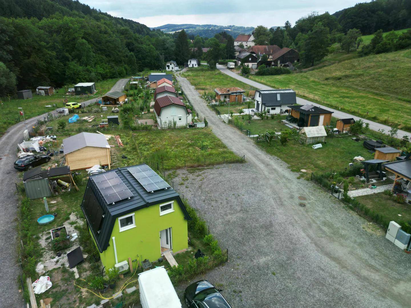 Luftaufnahme einer kleinen Siedlung mit verschiedenen Häusern, Schotterwegen und grüner Umgebung, einige Dächer mit Solarpaneelen.