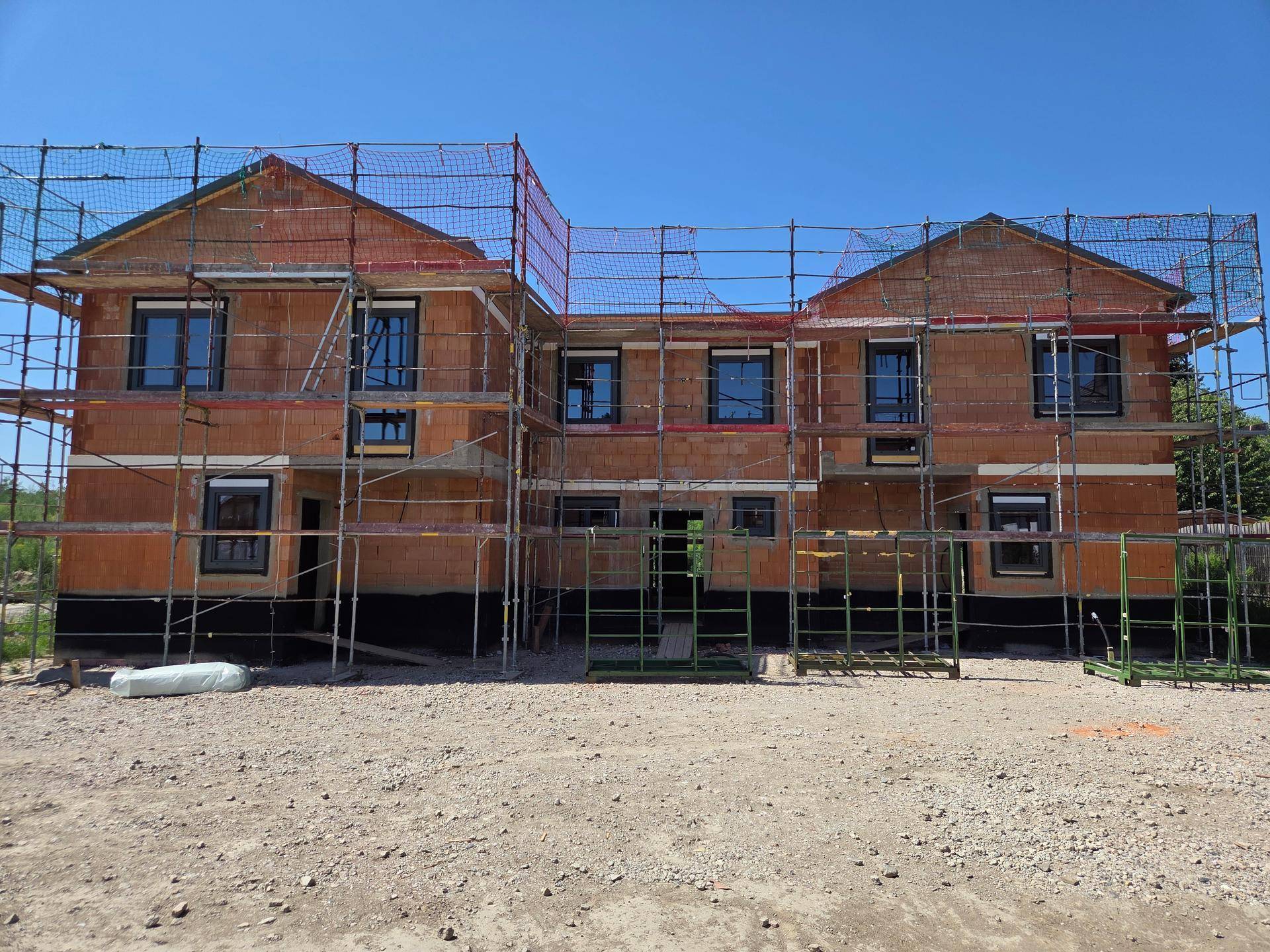 Neubau-Doppelhäuser im Rohbau mit Gerüst und Bauzaun unter blauem Himmel.