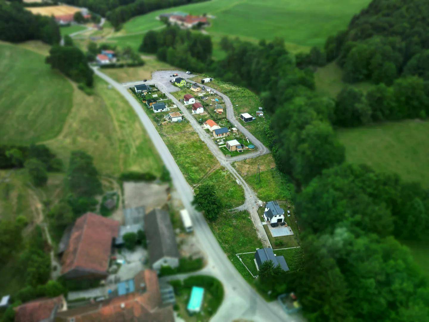 Weitläufige Luftaufnahme einer Siedlung, eingebettet in grüne Hügel und Wälder, mit einer Straße, die sich durch die Landschaft schlängelt.