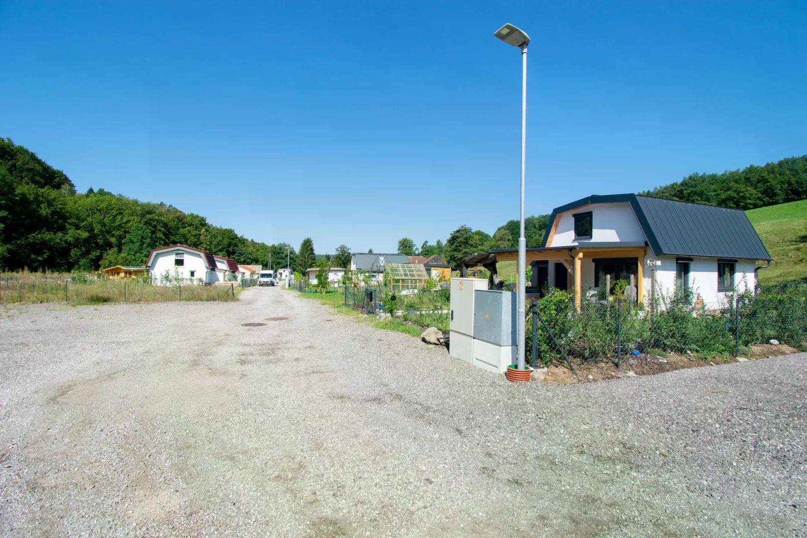 Sonniger Blick auf eine ländliche Siedlung mit Schotterweg, kleinen Häusern und einer Straßenlaterne vor grünen Hügeln.