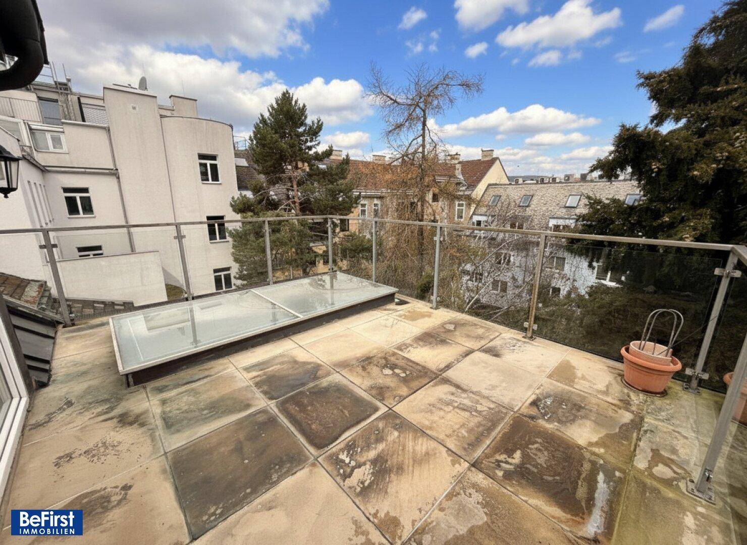 Große Dachterrasse mit Fliesenboden, Glasgeländer und Blick auf die umliegenden Gebäude.