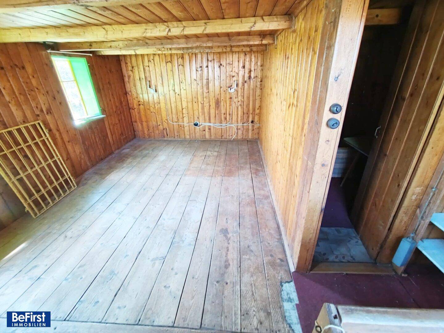 Leeres Zimmer mit Holzwänden, Holzboden und einem Fenster mit Blick ins Grüne.