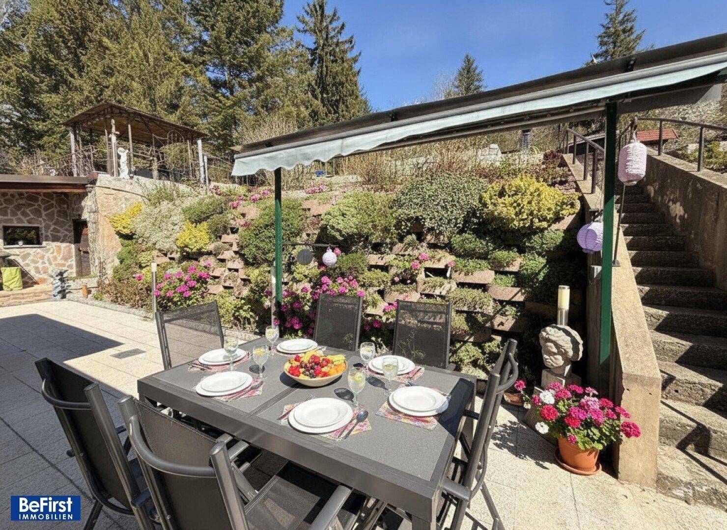 Gemütliche Terrasse mit Esstisch und Stühlen, umgeben von üppiger Bepflanzung und einer Markise.