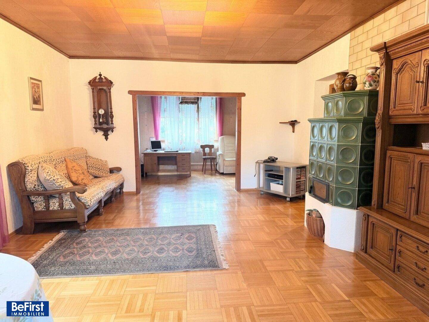 Helles Wohnzimmer mit Holzboden, Kachelofen und Zugang zu einem weiteren Raum.