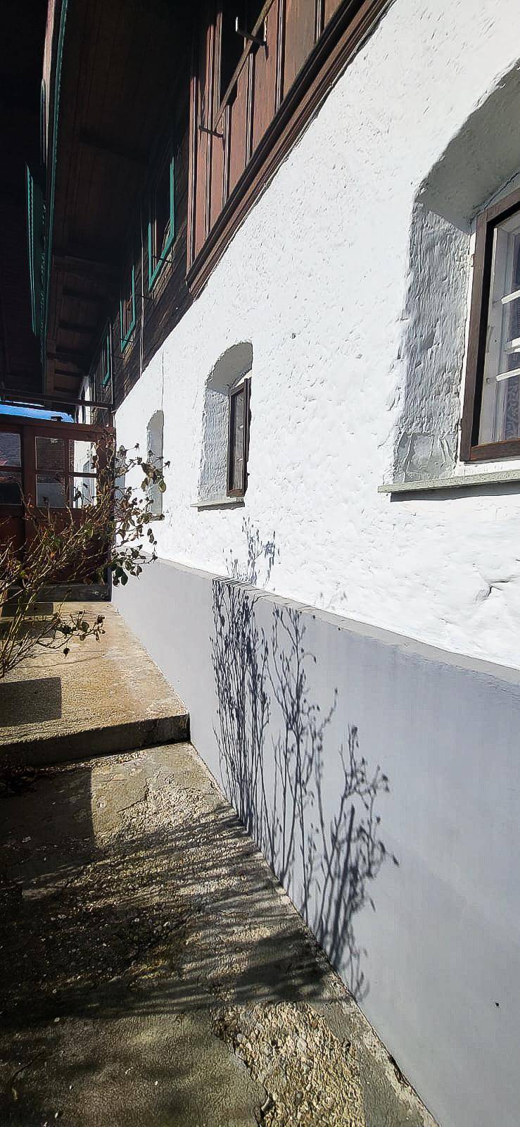 Traditionelle weiße Fassade eines Gebäudes mit kleinen Fenstern und einem Holzbalkon im Sonnenlicht.