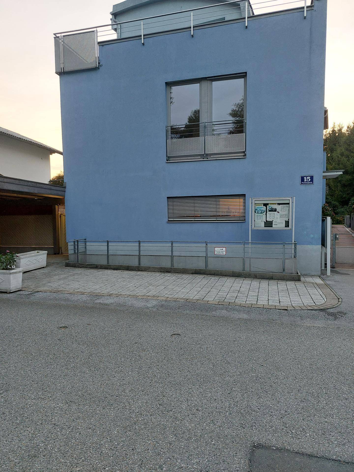 Moderne blaue Gebäudefassade mit großen Fenstern, kleinem Balkon und barrierefreier Rampe.