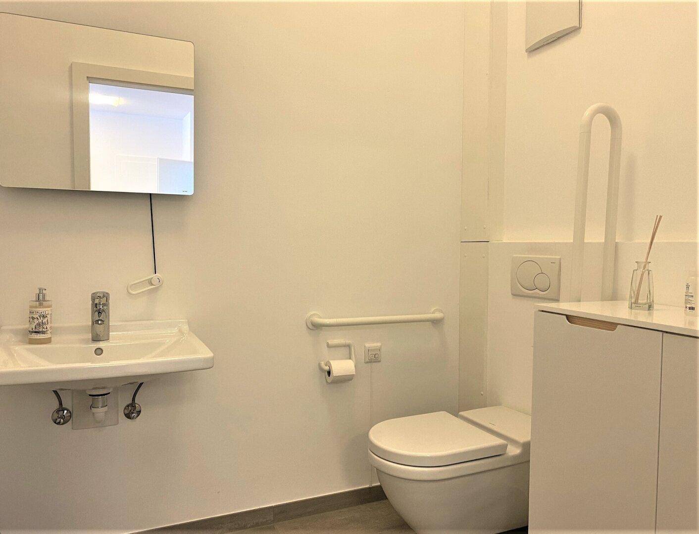 Modernes Badezimmer mit WC, Waschbecken und Haltegriffen für Barrierefreiheit, in hellen Farbtönen gehalten.