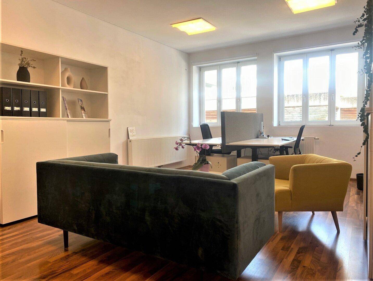 Geräumiges Büro mit dunklem Sofa, gelbem Sessel und Regalen für Ablage, gut beleuchtet durch Fenster.