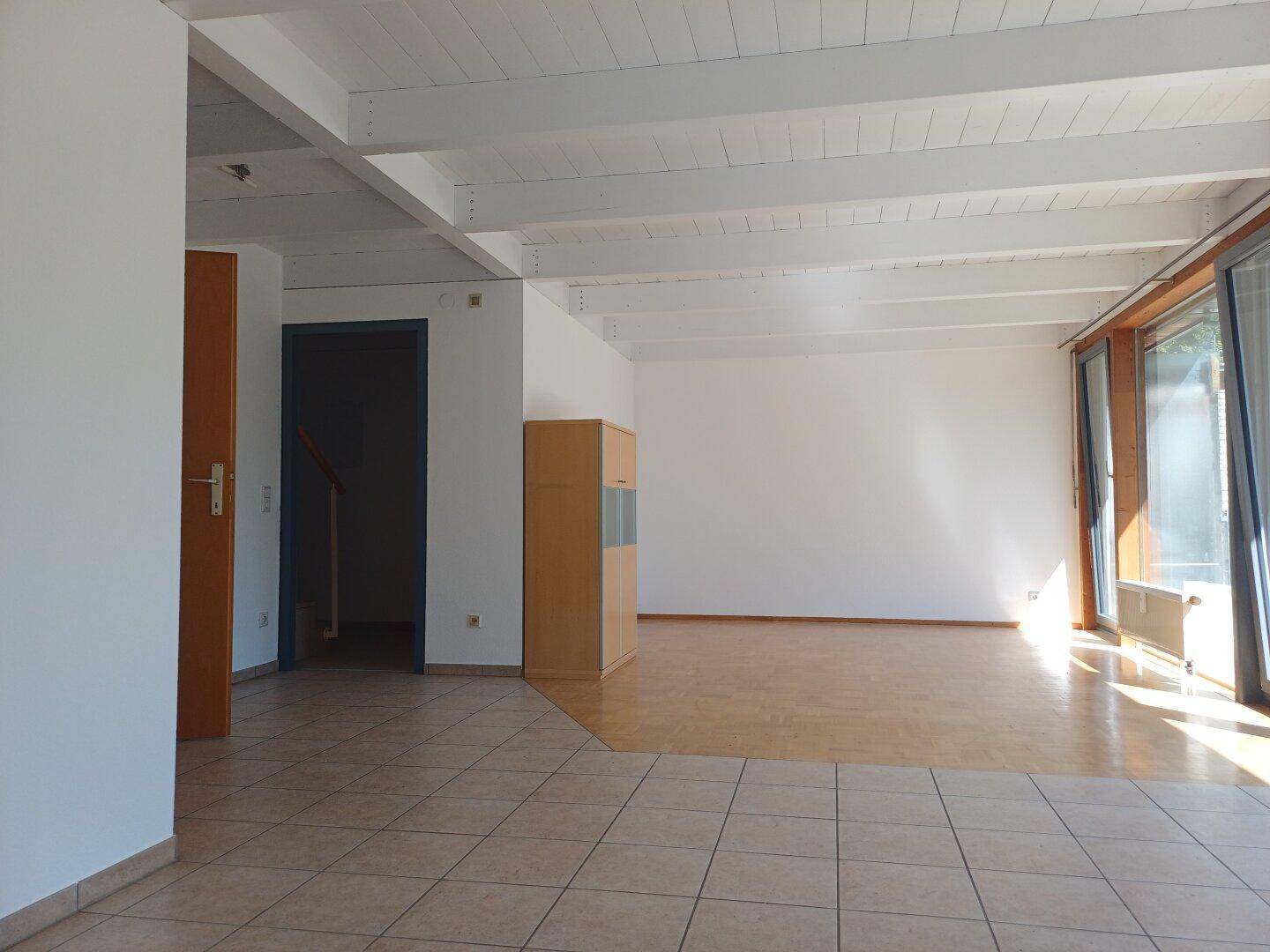 Lichtdurchfluteter Wohnbereich mit großen Fenstern, sichtbaren Holzbalken und kombiniertem Bodenbelag.