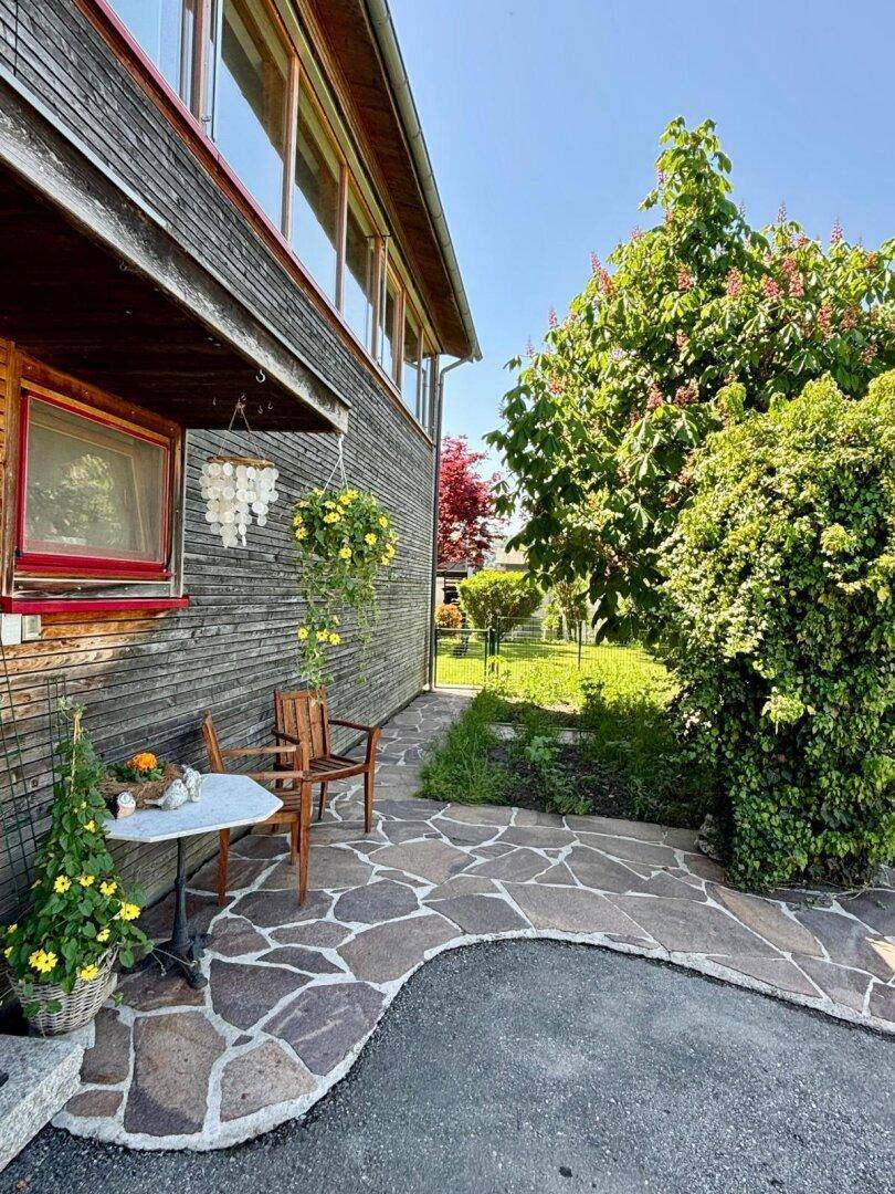 Kleiner, gepflasterter Sitzbereich neben dem Haus, umgeben von üppigem Grün und Bäumen.
