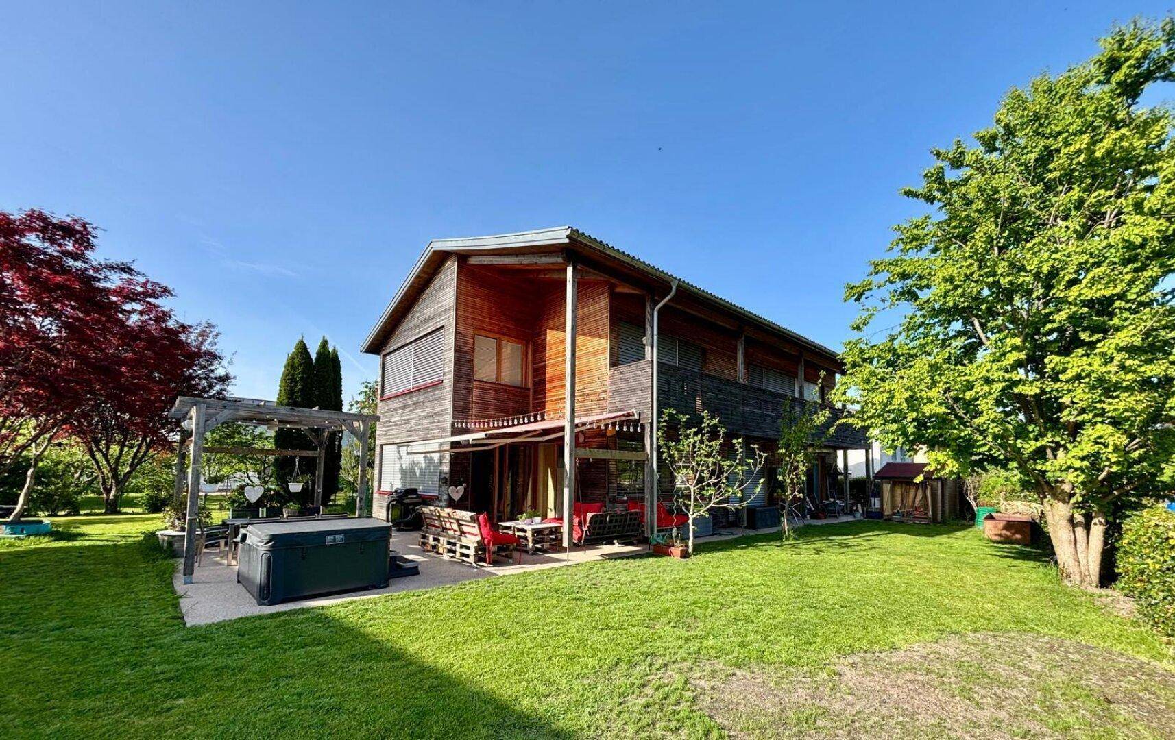 Weitläufiger Garten mit Holzhaus, großer Terrasse, Whirlpool und viel Grünfläche unter blauem Himmel.