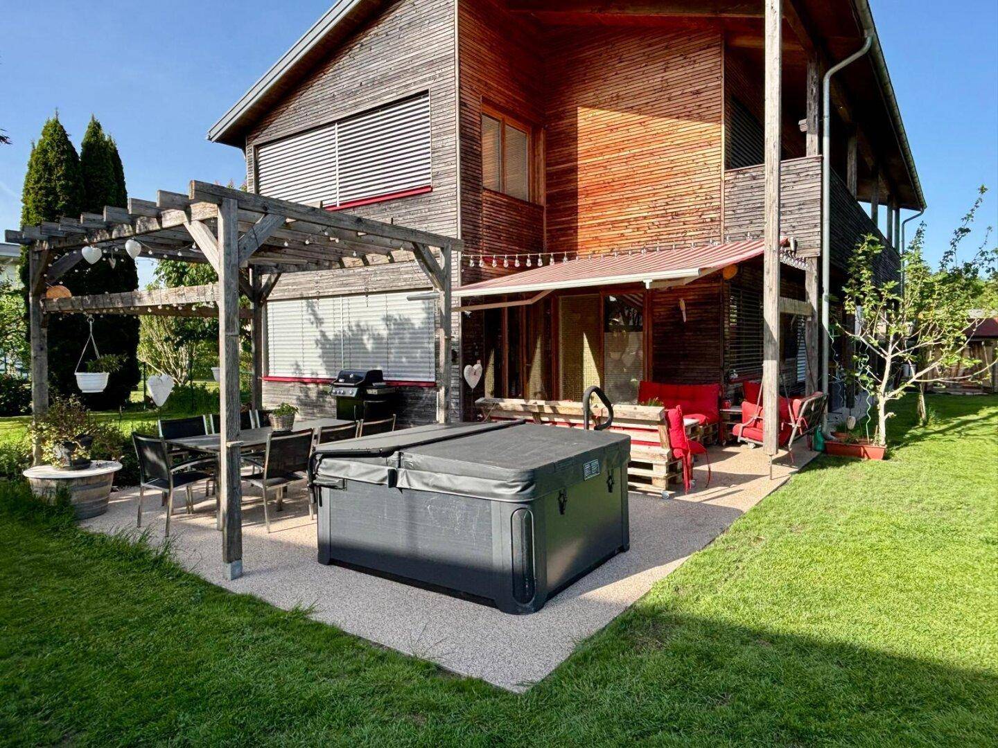 Sonniger Garten mit Holzhaus, Whirlpool und Essbereich unter einer Pergola, umgeben von Rasen.