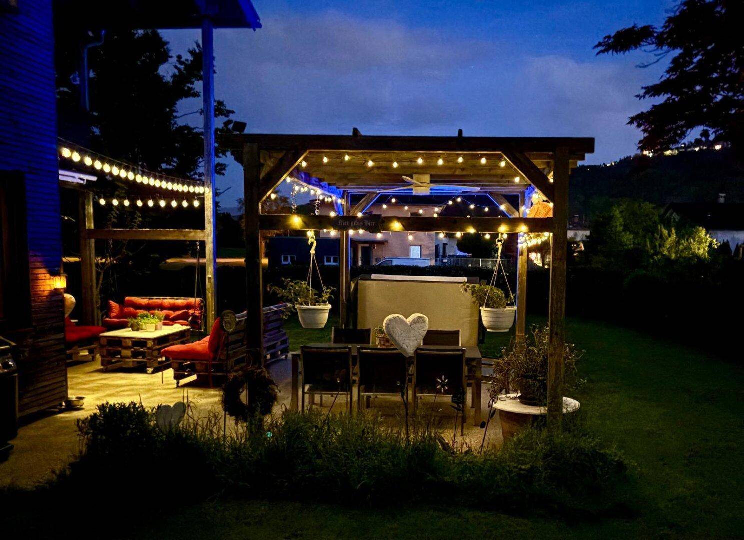 Stimmungsvoll beleuchteter Gartenbereich bei Nacht mit Pergola, Esstisch und Lichterketten.