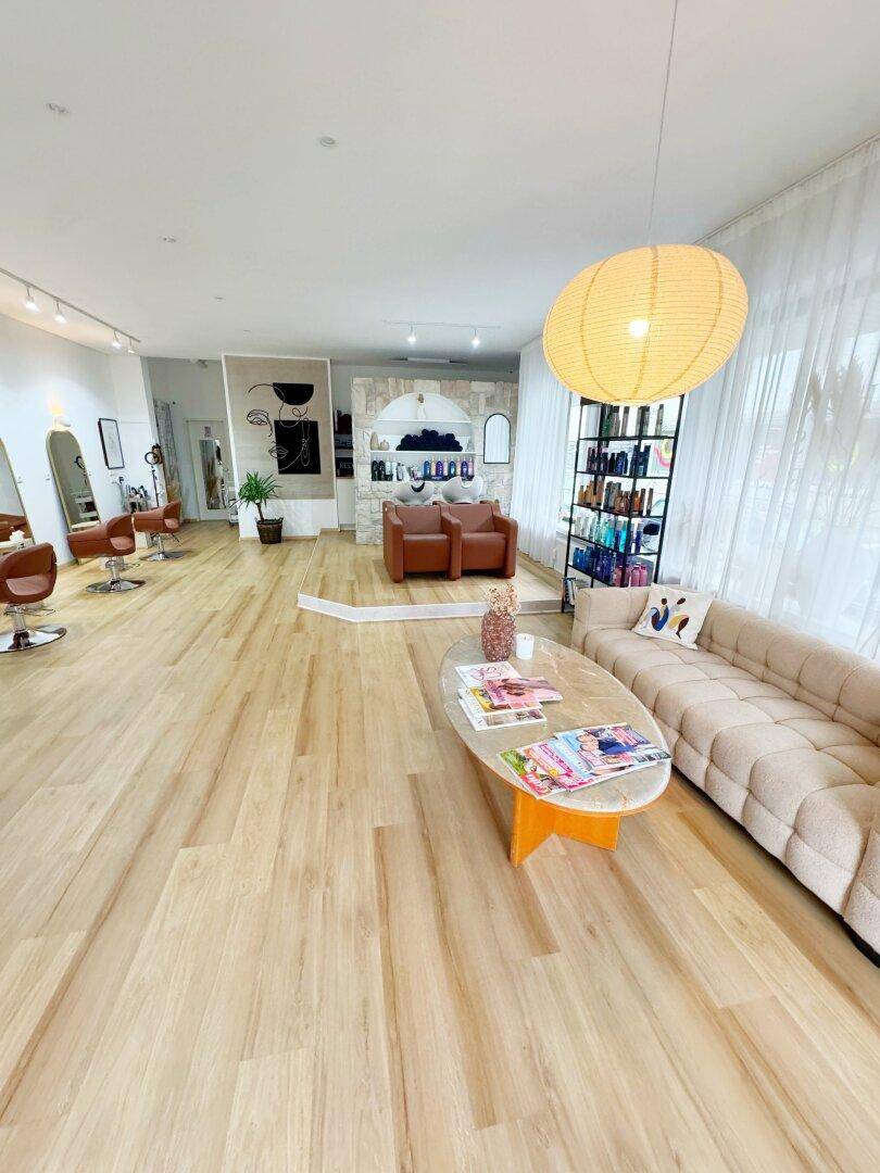 Geräumiger und heller Friseursalon mit modernen Stylingstühlen, Holzboden und gemütlicher Sitzecke.