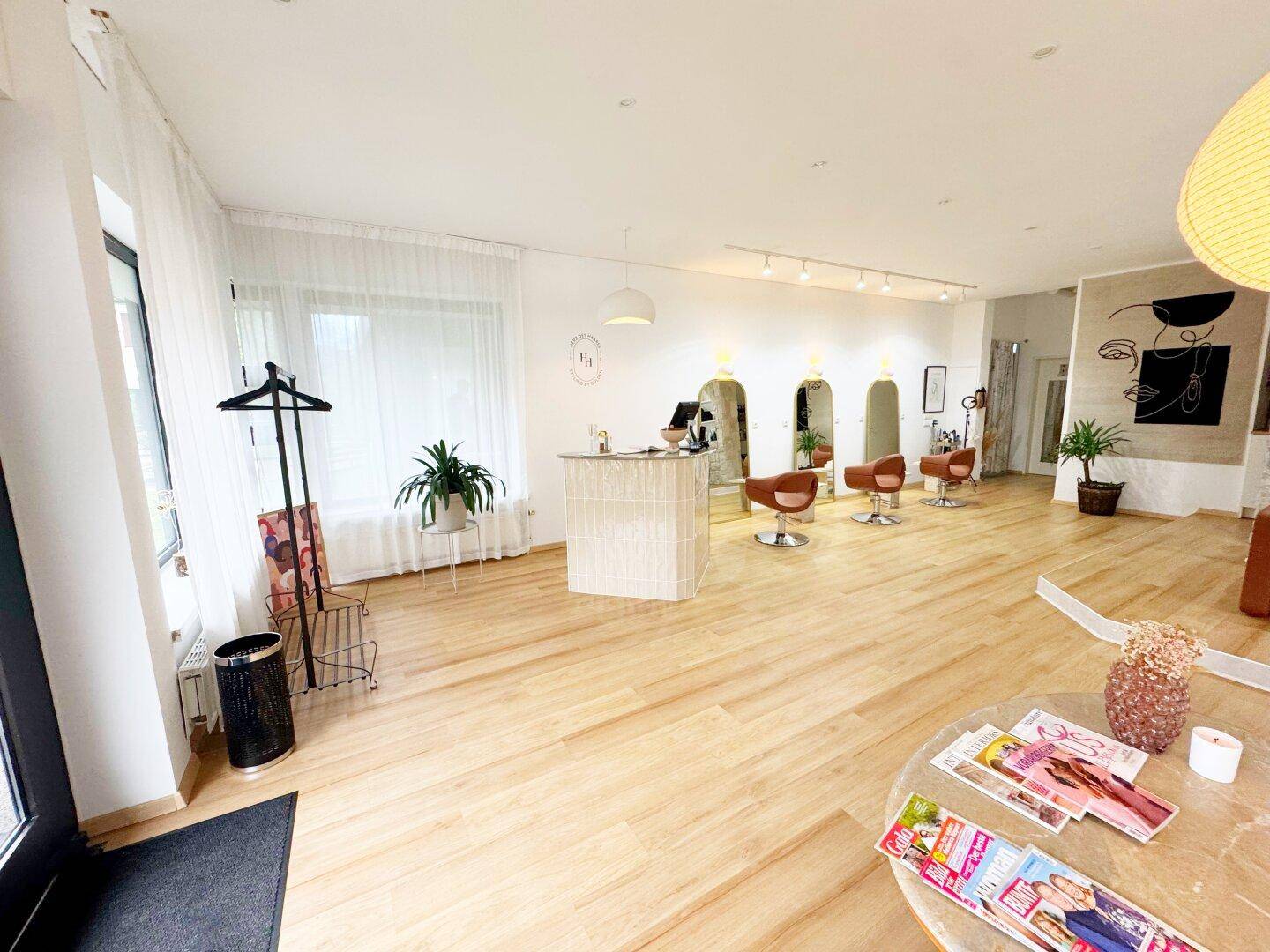 Empfangsbereich des Friseursalons mit modernem Tresen, Stylingstühlen und viel Tageslicht durch große Fenster.