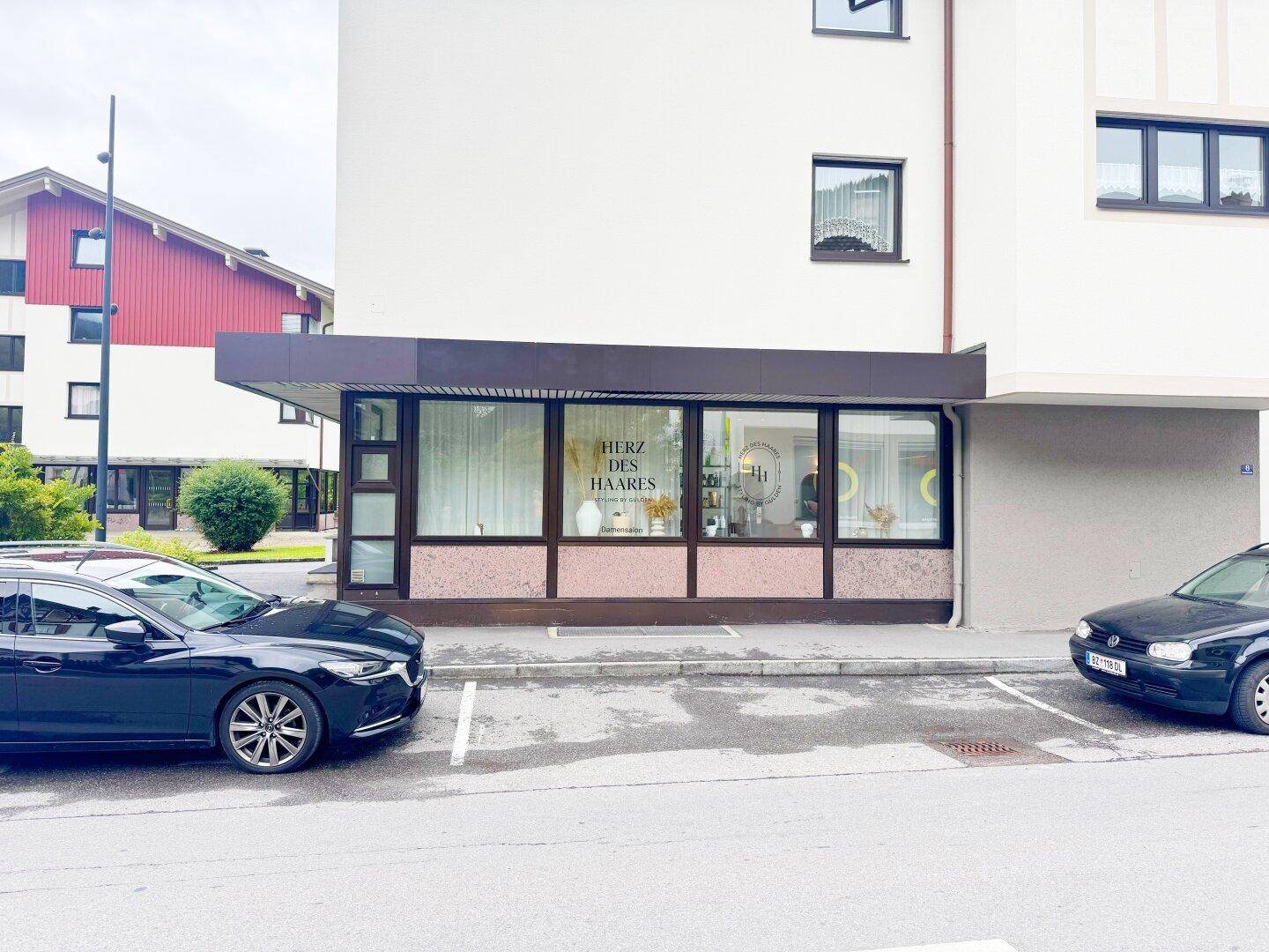 Ansicht der Geschäftsfassade mit großen Fenstern und davor geparkten Autos an der Straße.