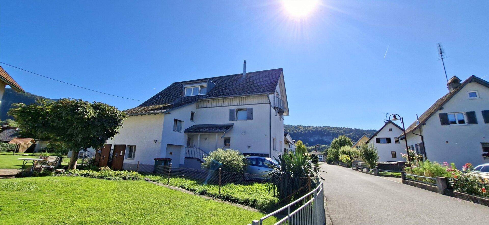Vorderansicht des weißen Wohnhauses mit Eingangsbereich und grünem Vorgarten unter strahlendem Sonnenschein.