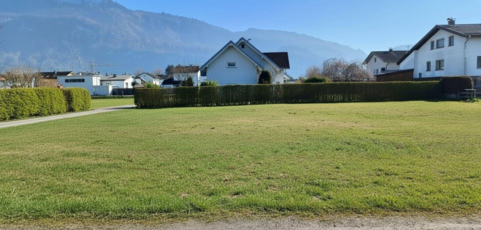 Weitläufige Rasenfläche mit Wohnhäusern und Bergpanorama im Hintergrund bei klarem Wetter.