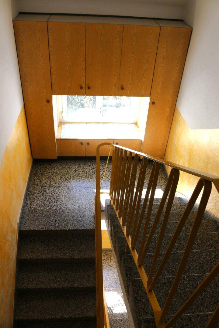 Treppenhaus mit gelbem Geländer, Terrazzoboden und Einbauschränken unter dem Fenster.