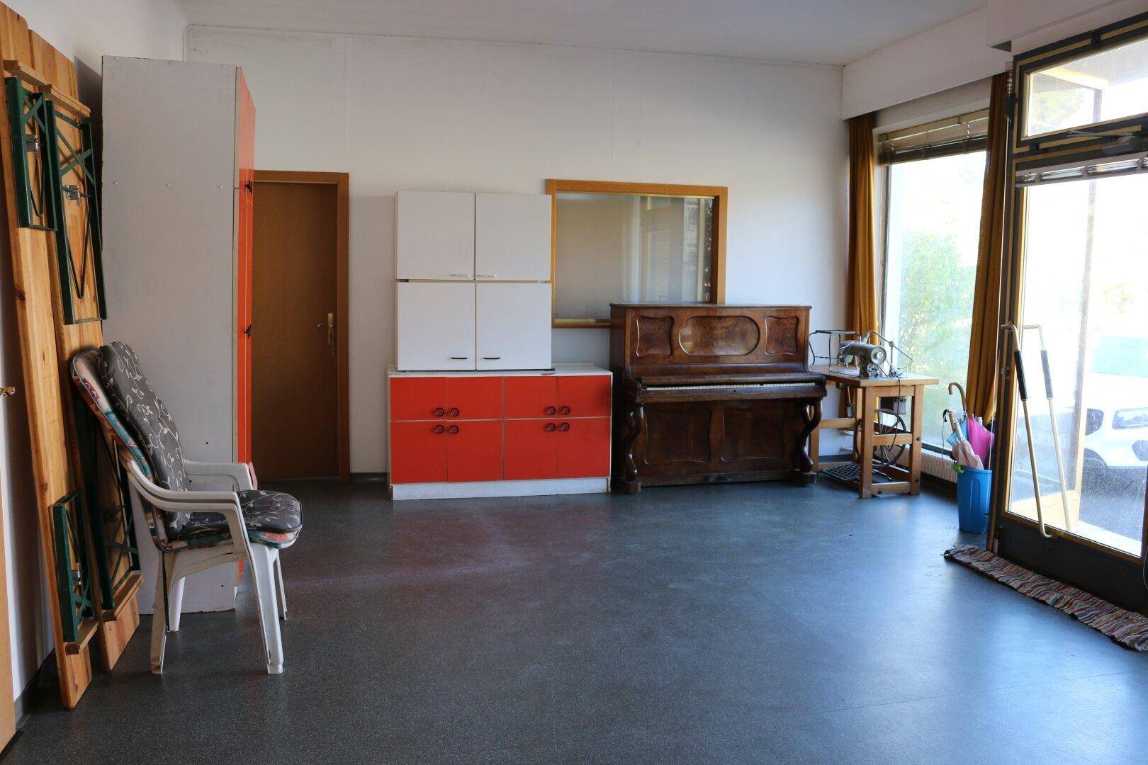 Großer, heller Raum mit dunklem Boden, großen Fenstern und verschiedenen Möbelstücken.