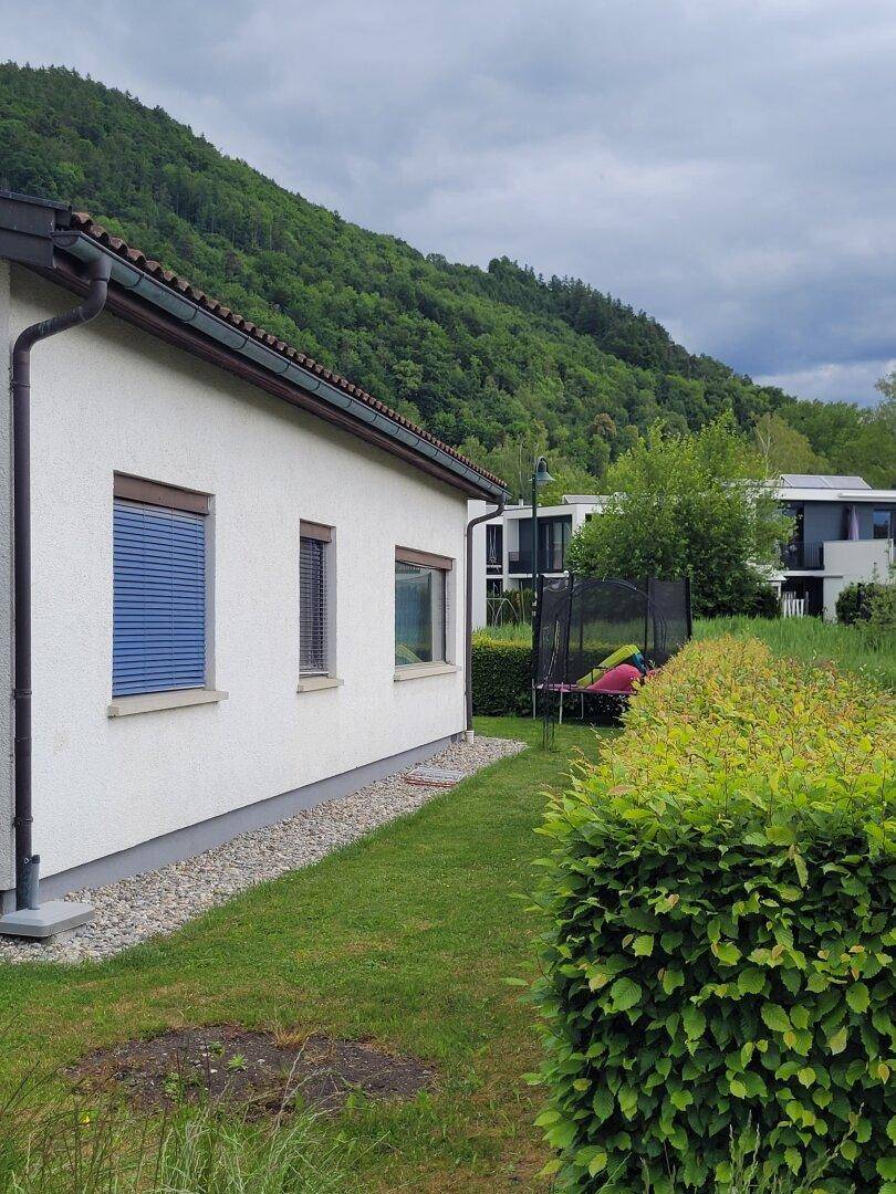 Seitenansicht des Hauses mit Garten, Trampolin und Blick auf die bewaldeten Berge unter bewölktem Himmel.