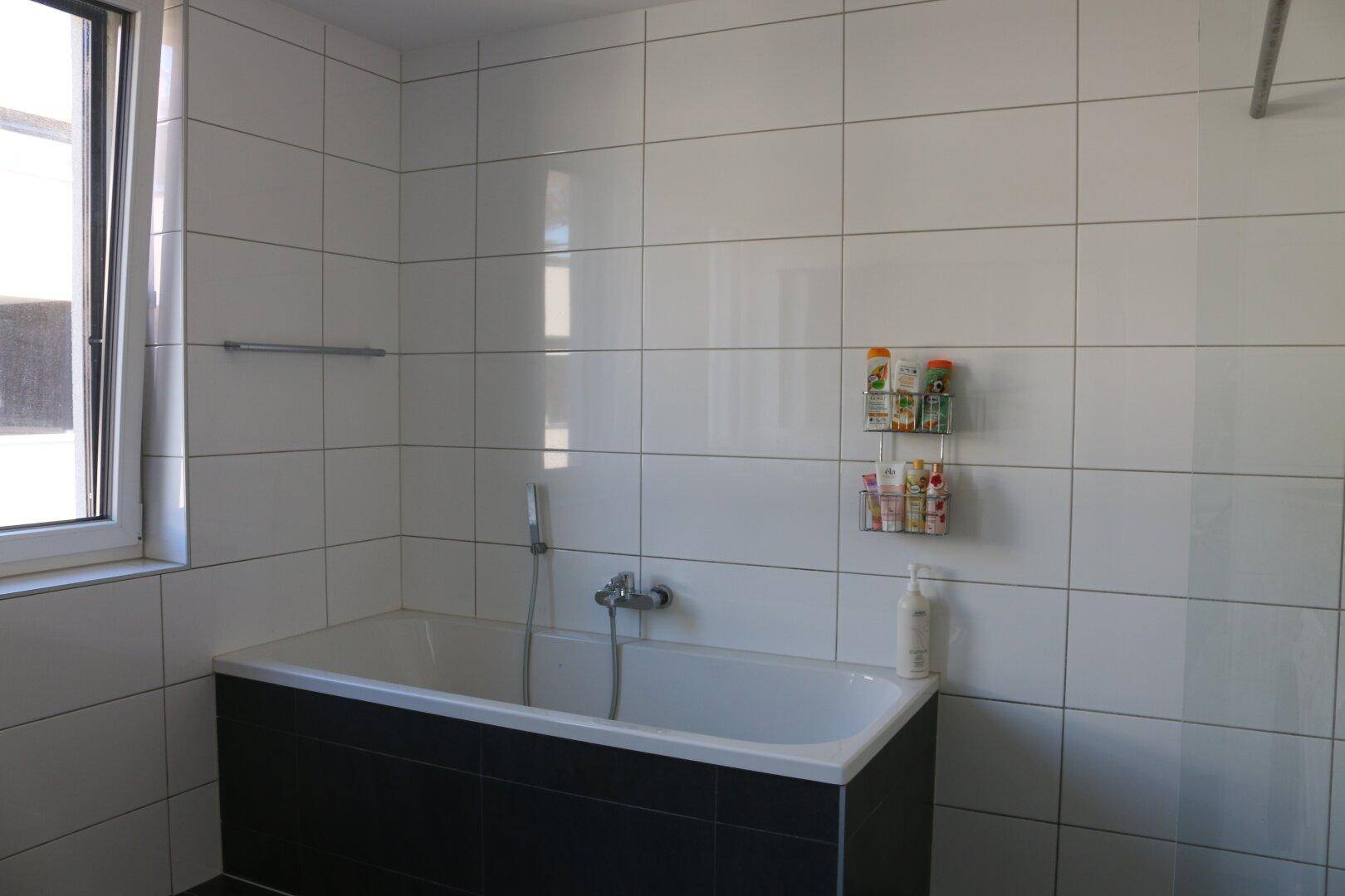 Helles Badezimmer mit Badewanne, weißen Wandfliesen und Fenster für natürliche Belüftung.