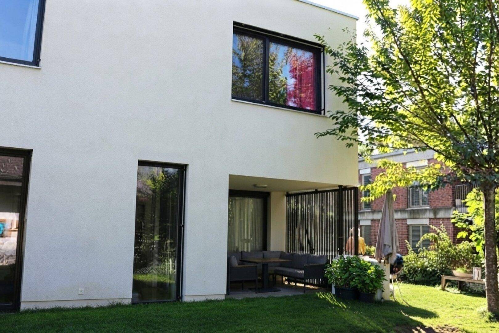 Moderne Hausfassade mit großen Fenstern und Zugang zu einer überdachten Terrasse.