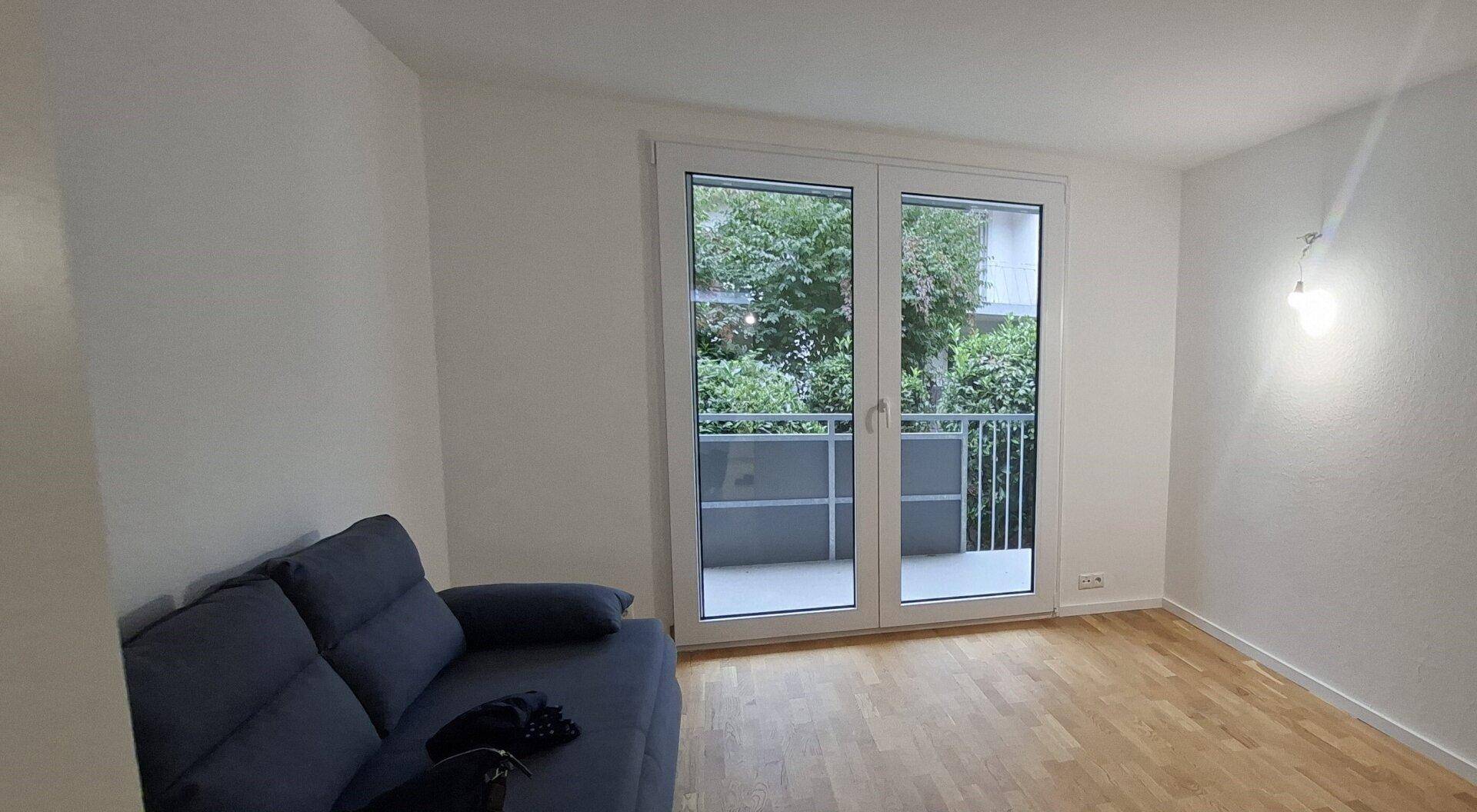 Helles Wohnzimmer mit Holzboden, einem Sofa und direktem Zugang zu einem Balkon mit Grünblick.