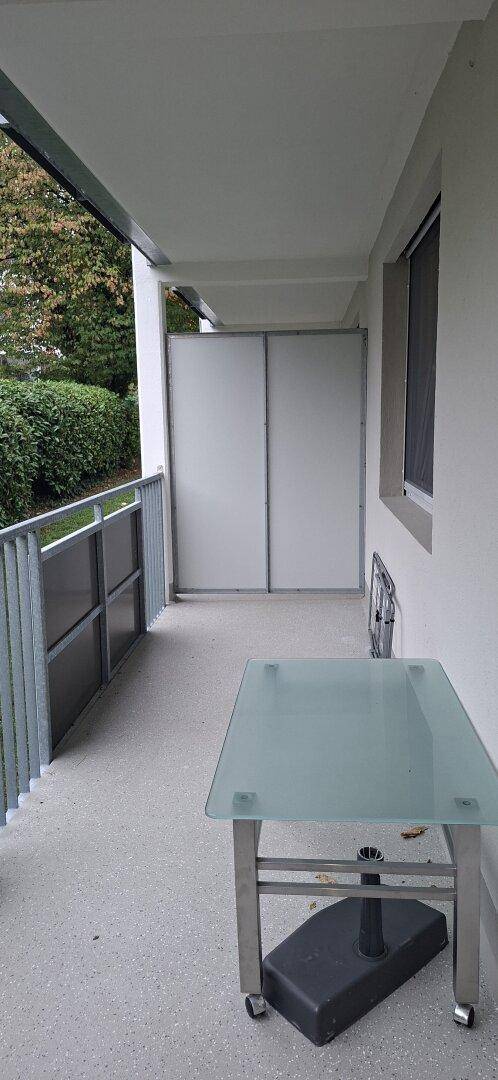 Überdachter Balkon mit Glasgeländer, einem kleinen Tisch und einem angenehmen Blick ins Grüne.
