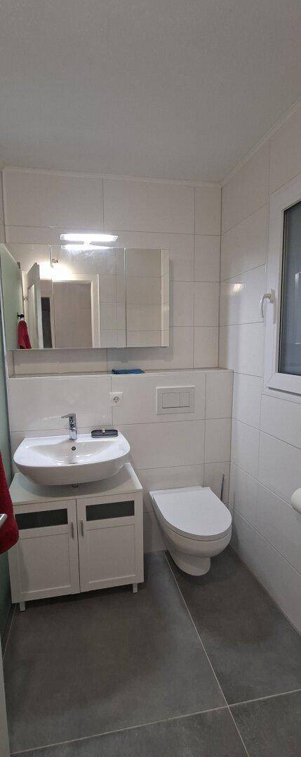 Modernes Badezimmer mit weißen Fliesen, Einzelwaschbecken, Toilette und einem Fenster für Tageslicht.