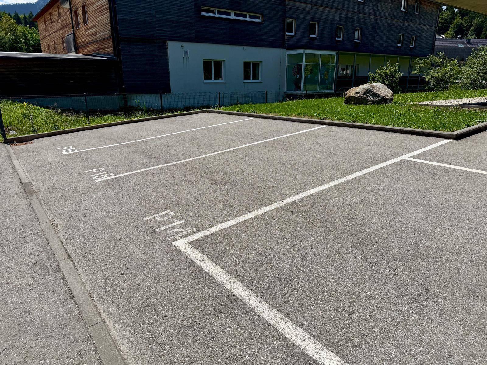 Mehrere markierte Außenparkplätze vor einem Gebäude mit Holzverkleidung und umgebender Grünfläche.