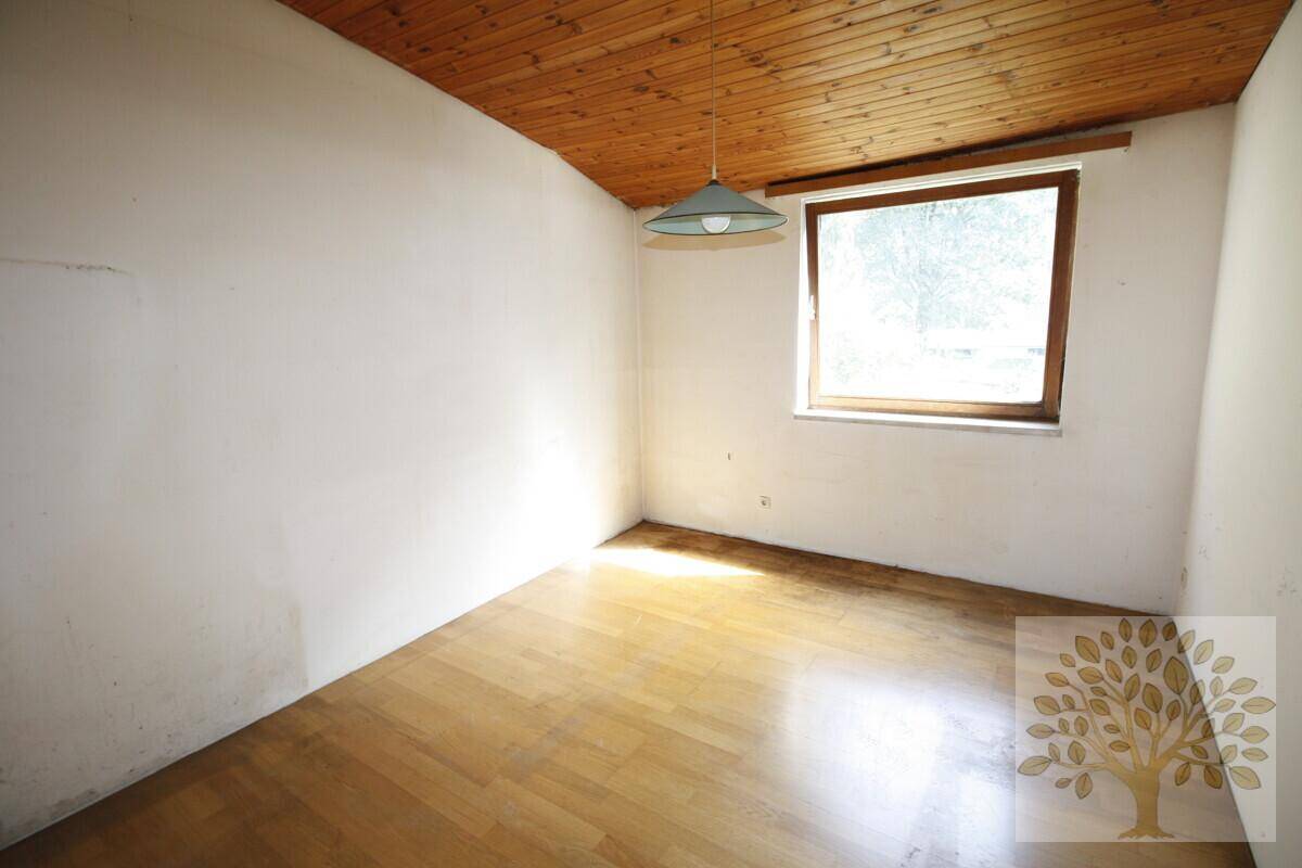 Helles Zimmer mit Holzparkettboden, Holzdecke und einem Fenster, das den Blick ins Grüne freigibt.