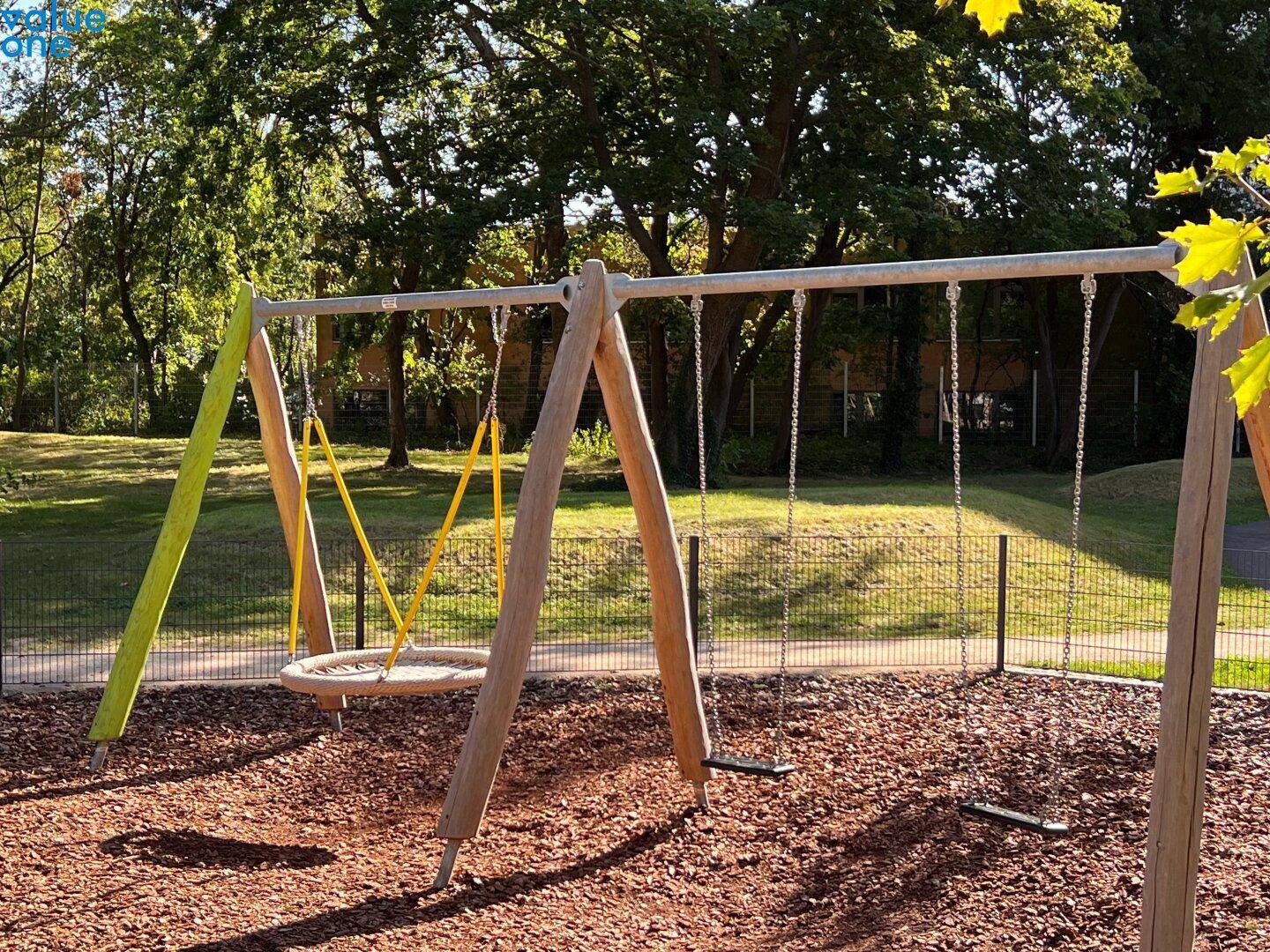 Kinderspielplatz mit Schaukeln und Rindenmulchboden, umgeben von Bäumen.