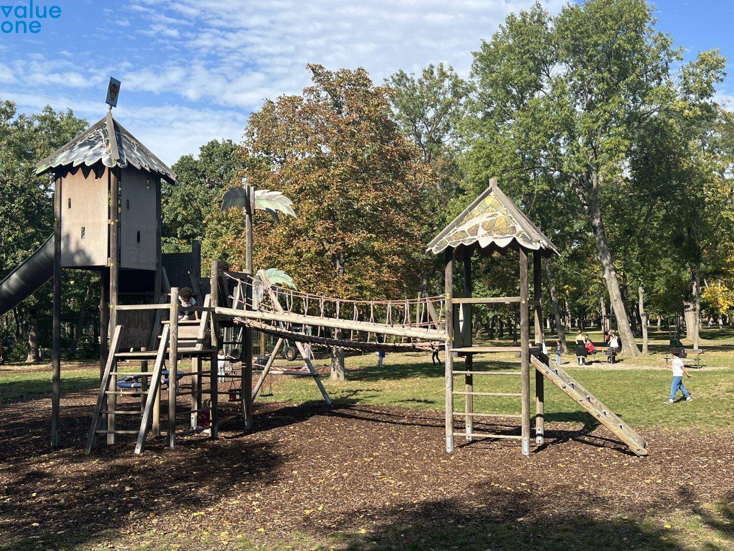 Kinderspielplatz mit Holzspielgeräten und Seilbrücke in einer grünen Parkanlage.