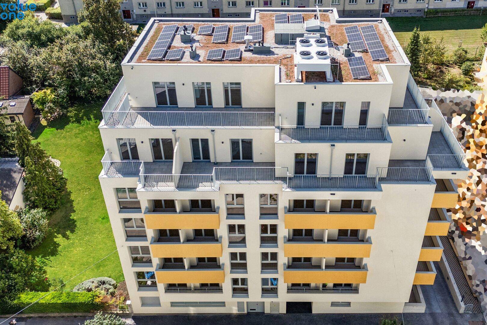 Vogelperspektive eines modernen Wohnkomplexes mit Solarpaneelen auf dem Flachdach und angrenzenden Grünflächen.