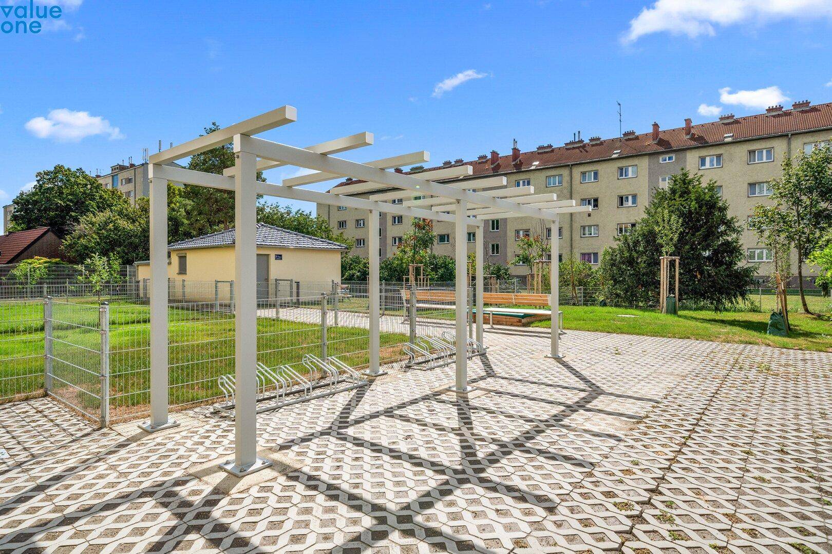 Überdachter Fahrradabstellplatz auf einem gepflasterten Bereich mit Bänken und Grünflächen.