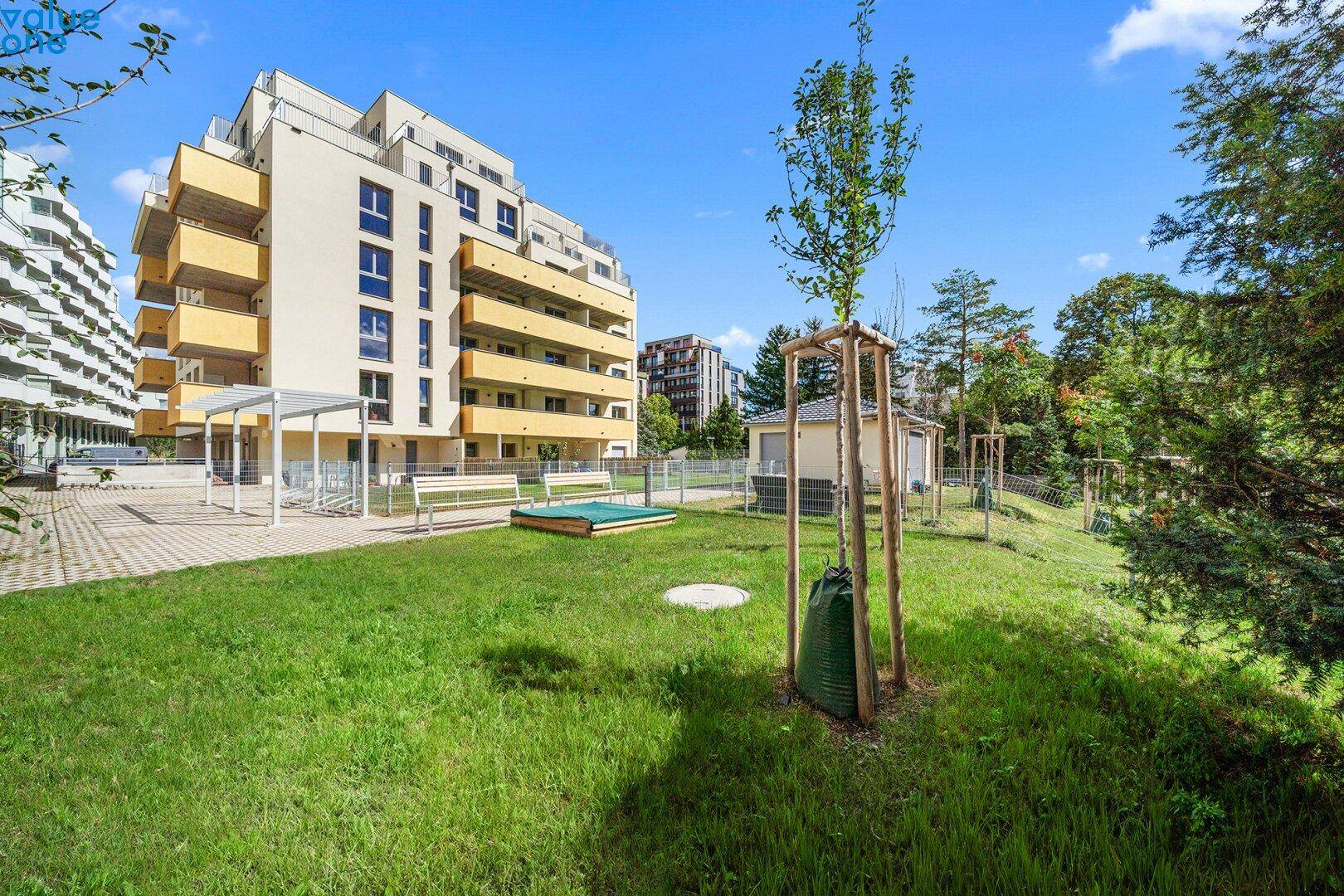 Gepflegte Grünfläche mit Sandkasten, Bänken und jungen Bäumen vor einem modernen Wohnhaus.