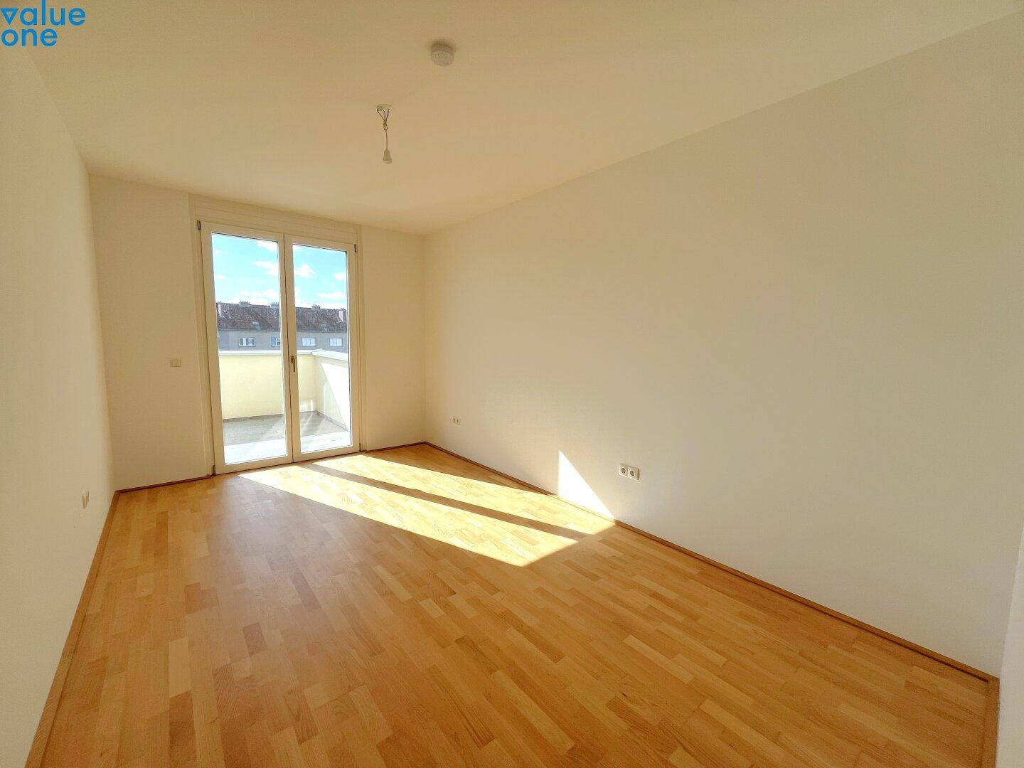 Leeres Zimmer mit Parkettboden und großen Fenstern, die viel Tageslicht hereinlassen.