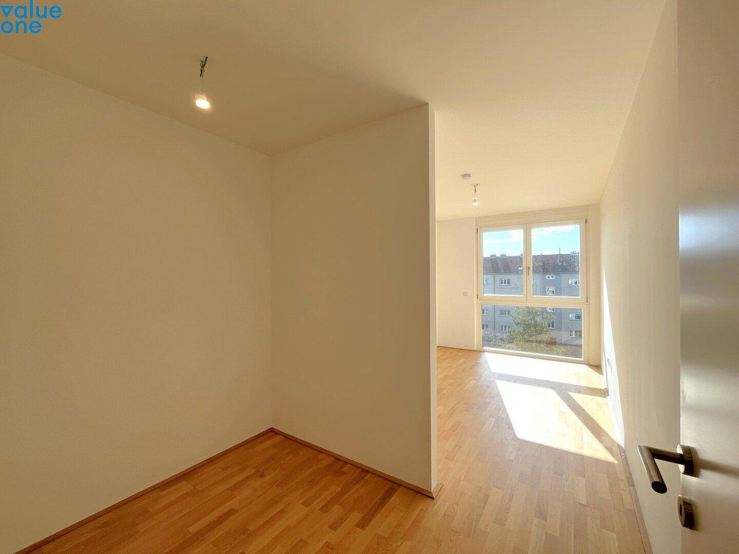 Helles, leeres Zimmer mit Holzboden und großem Fenster, ideal als Schlaf- oder Arbeitszimmer.