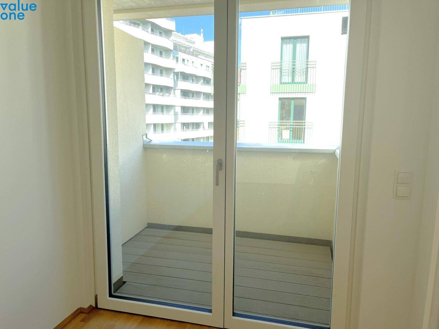 Blick vom Innenraum auf einen leeren Balkon mit Holzboden und Blick auf umliegende Gebäude.