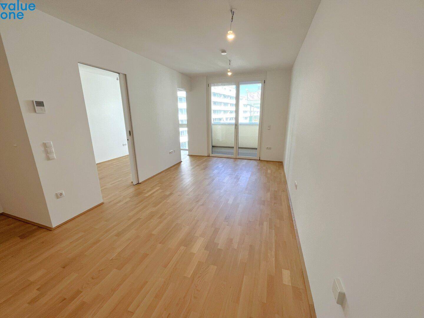 Geräumiges, leeres Wohnzimmer mit Holzboden und großen Fenstern, die viel Licht hereinlassen.