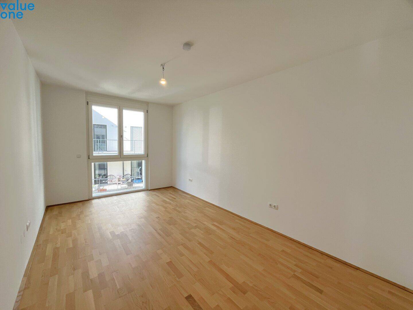 Geräumiger, leerer Raum mit Holzboden und großen Fenstern, die viel Tageslicht hereinlassen.