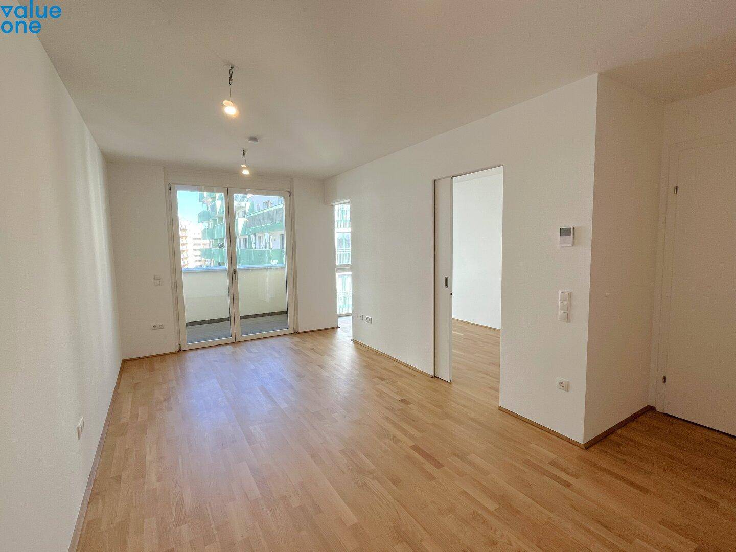 Geräumiges, leeres Wohnzimmer mit hellem Parkettboden und Zugang zum Balkon.