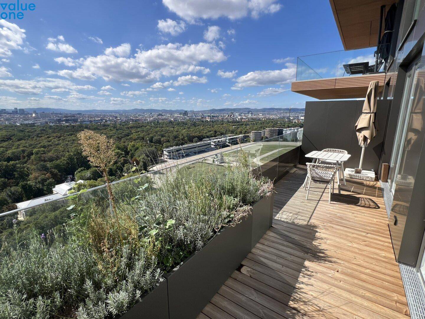 Geräumiger Balkon mit Holzboden, Pflanzen und weitem Blick über die grüne Stadtlandschaft.