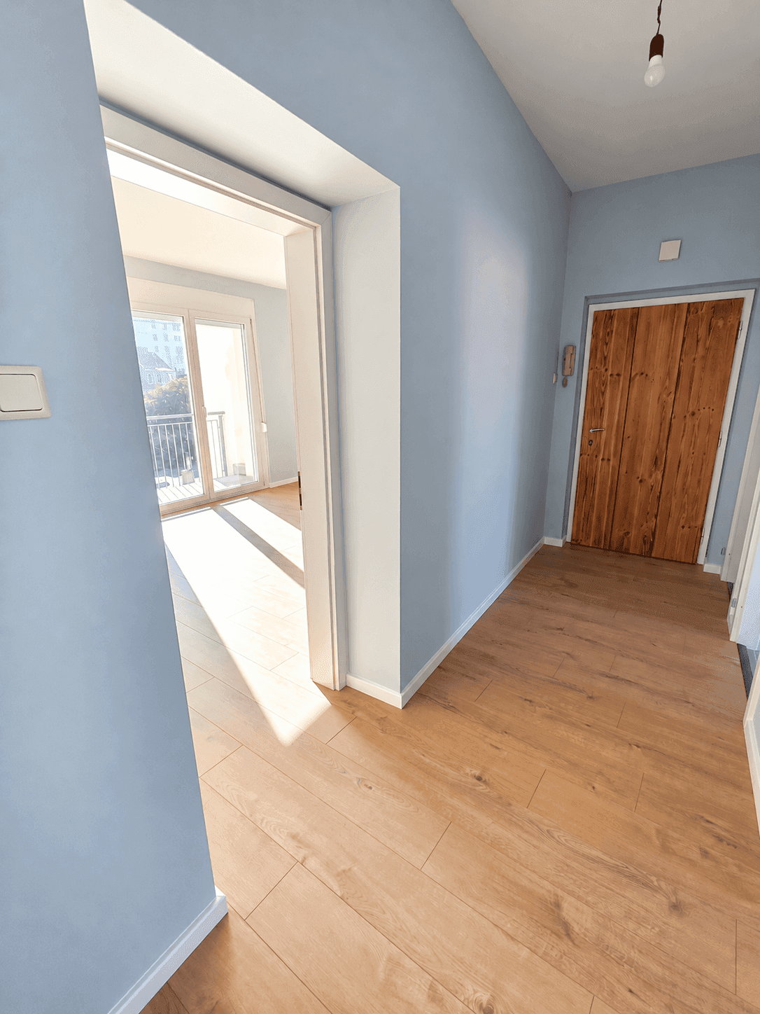 Heller Flur mit Laminatboden und hellblauen Wänden, der einen Blick auf den sonnigen Balkon freigibt.